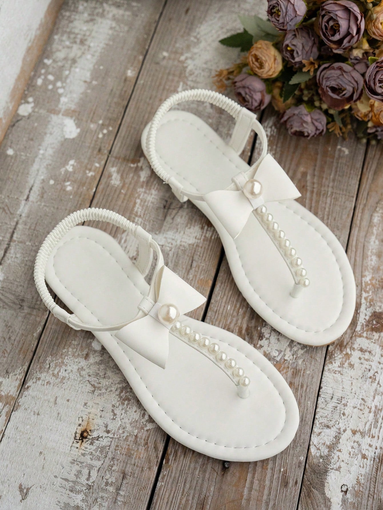 Beige women's flat thong Roman-style sandals with toe post, rhinestone and bow decoration, and buckle detail.
