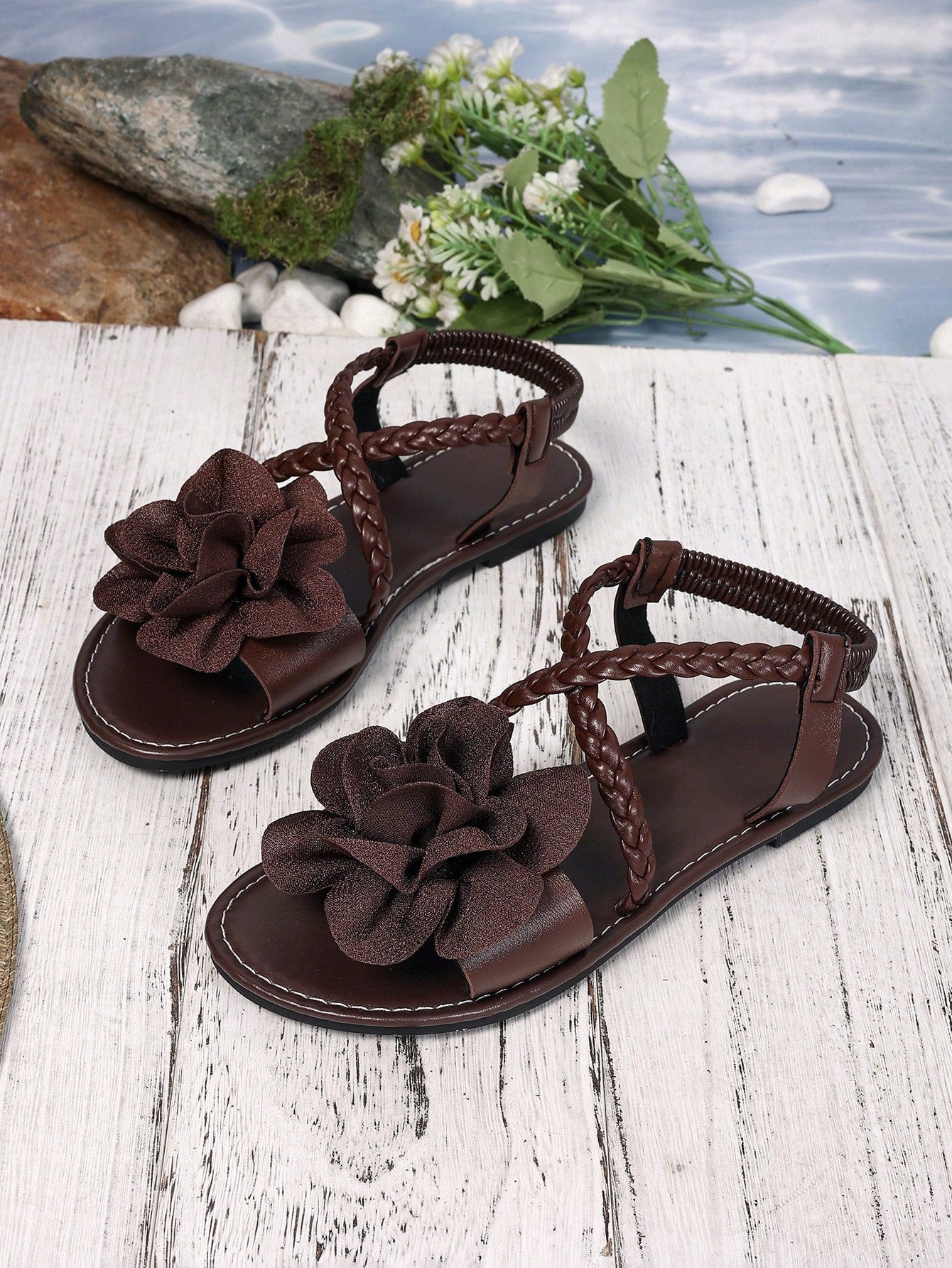 Women's open-toe flat slide sandals with crossed woven-look straps, floral appliqué detail, and buckle accent, shown in a casual beach style.