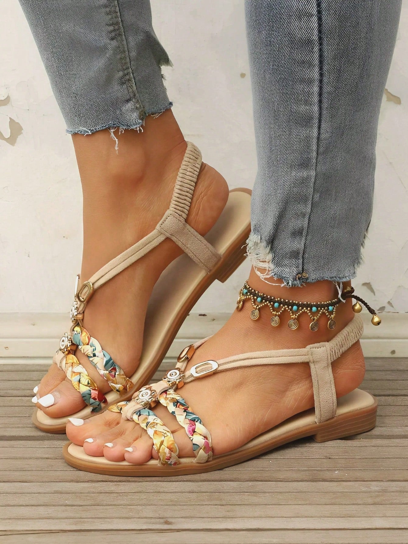 Women's beige flat gladiator sandals with elastic braided straps, floral decoration, rhinestone detail, and open toe, suitable for beach and vacation wear.