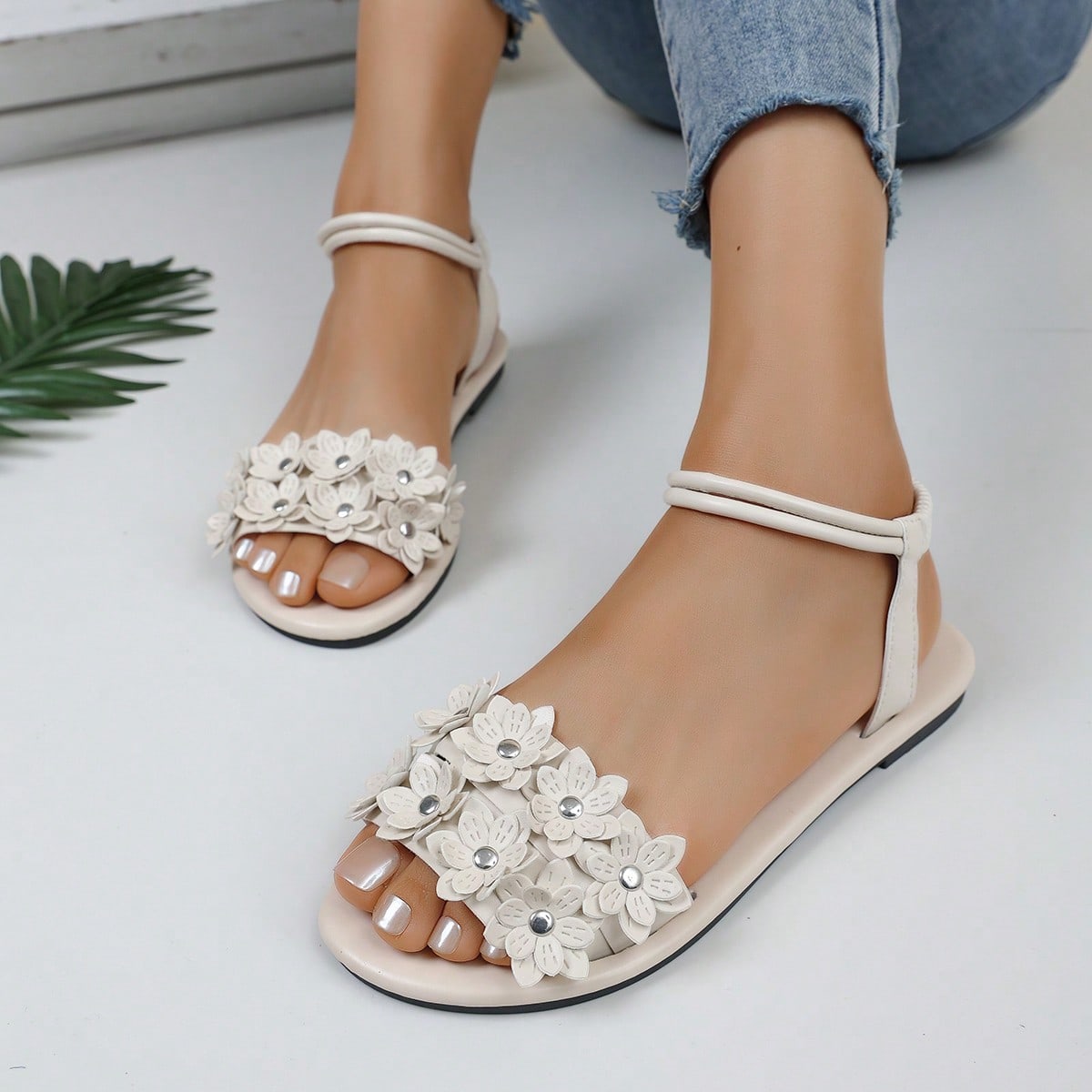 Women's beige flat sandals with stitched flower petal detail, open toe front, and adjustable ankle strap for casual warm-weather and beach wear.