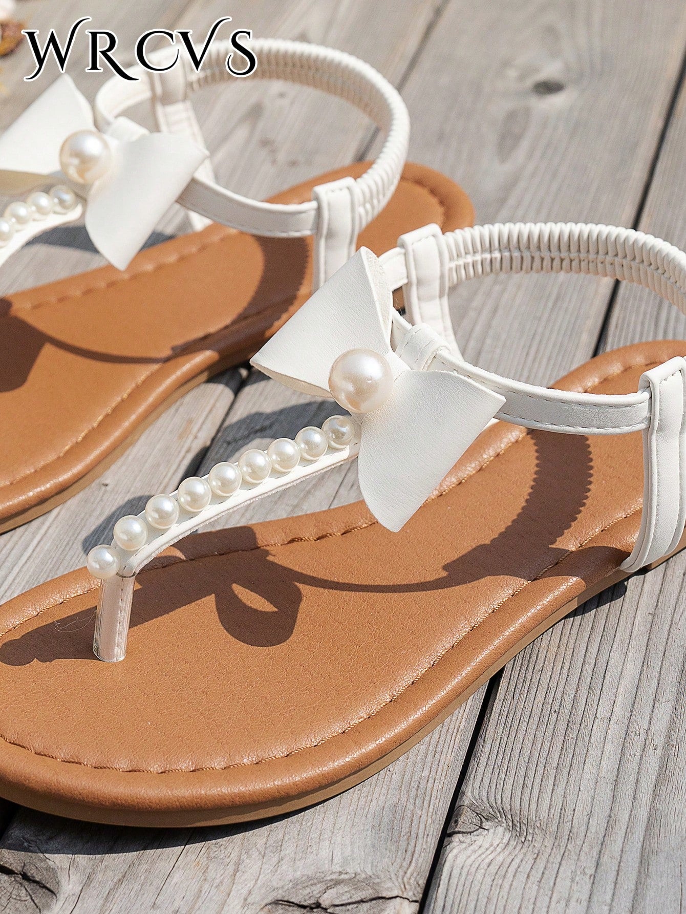 Beige women's flat thong Roman-style sandals with toe post, rhinestone and bow decoration, and buckle detail.